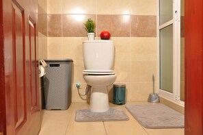 A bathroom with a toilet, a trash can, a small plant on the toilet lid, a red apple on the lid, and a window with a shower curtain.