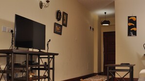 A living room with a wooden floor, a television on a stand, a fluffy rug, and a door in the background. - Sherry Homes - Fahari I (Nairobi)