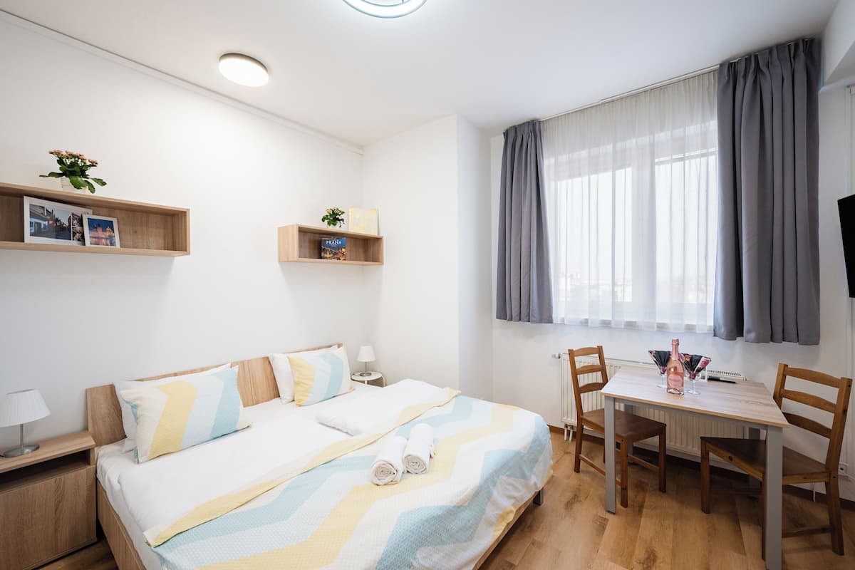 A bedroom with a bed, bedside tables, a desk with chairs, a window with curtains, and a wall-mounted TV.
