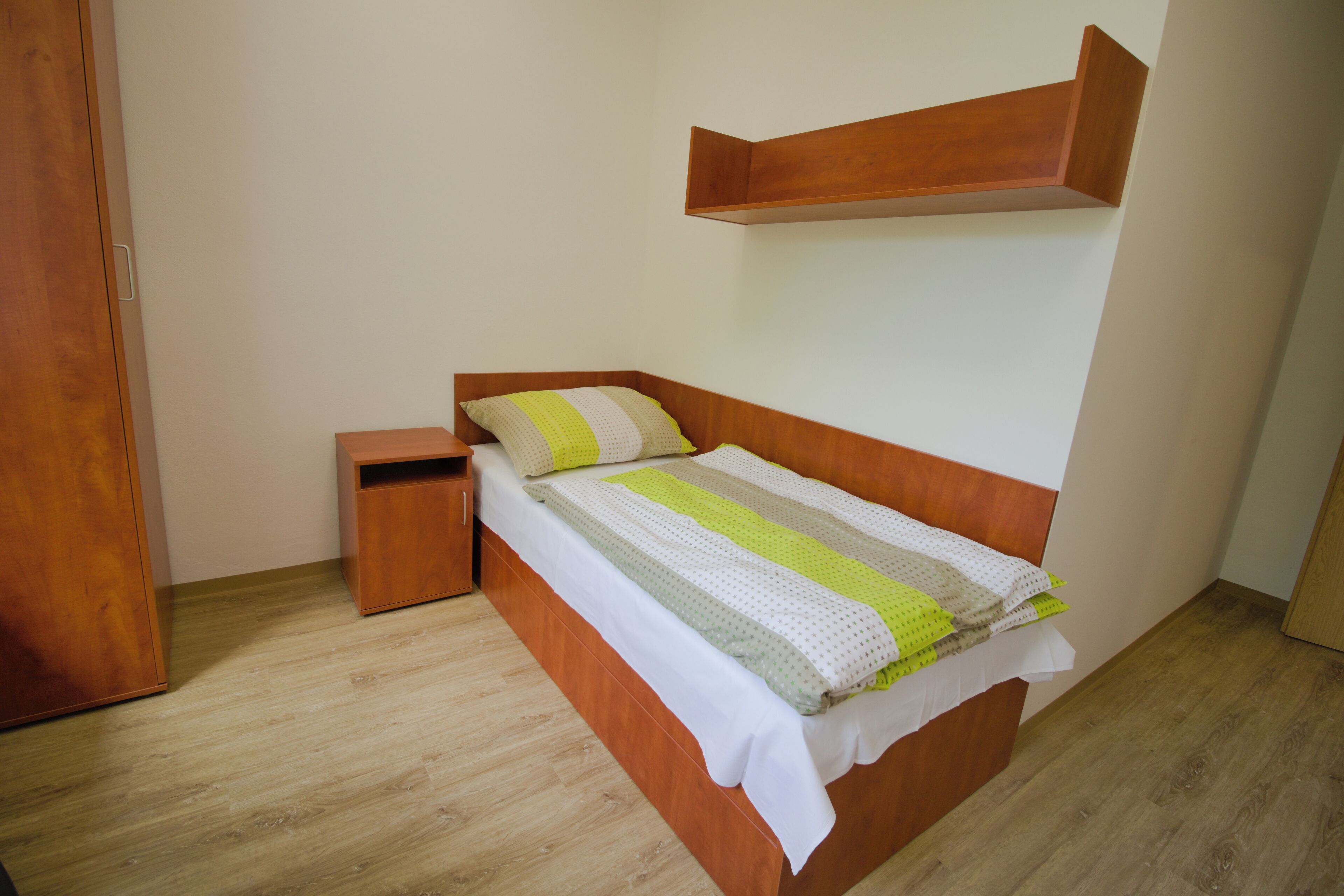 A bedroom with a wooden bed, a nightstand, a wooden wardrobe, and a shelf above the bed.