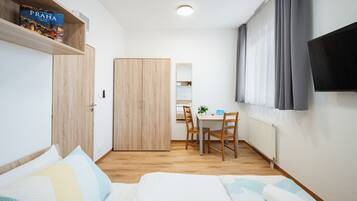 A bedroom with a bed, a desk, a chair, a TV, a wardrobe, and a shelf with a book.