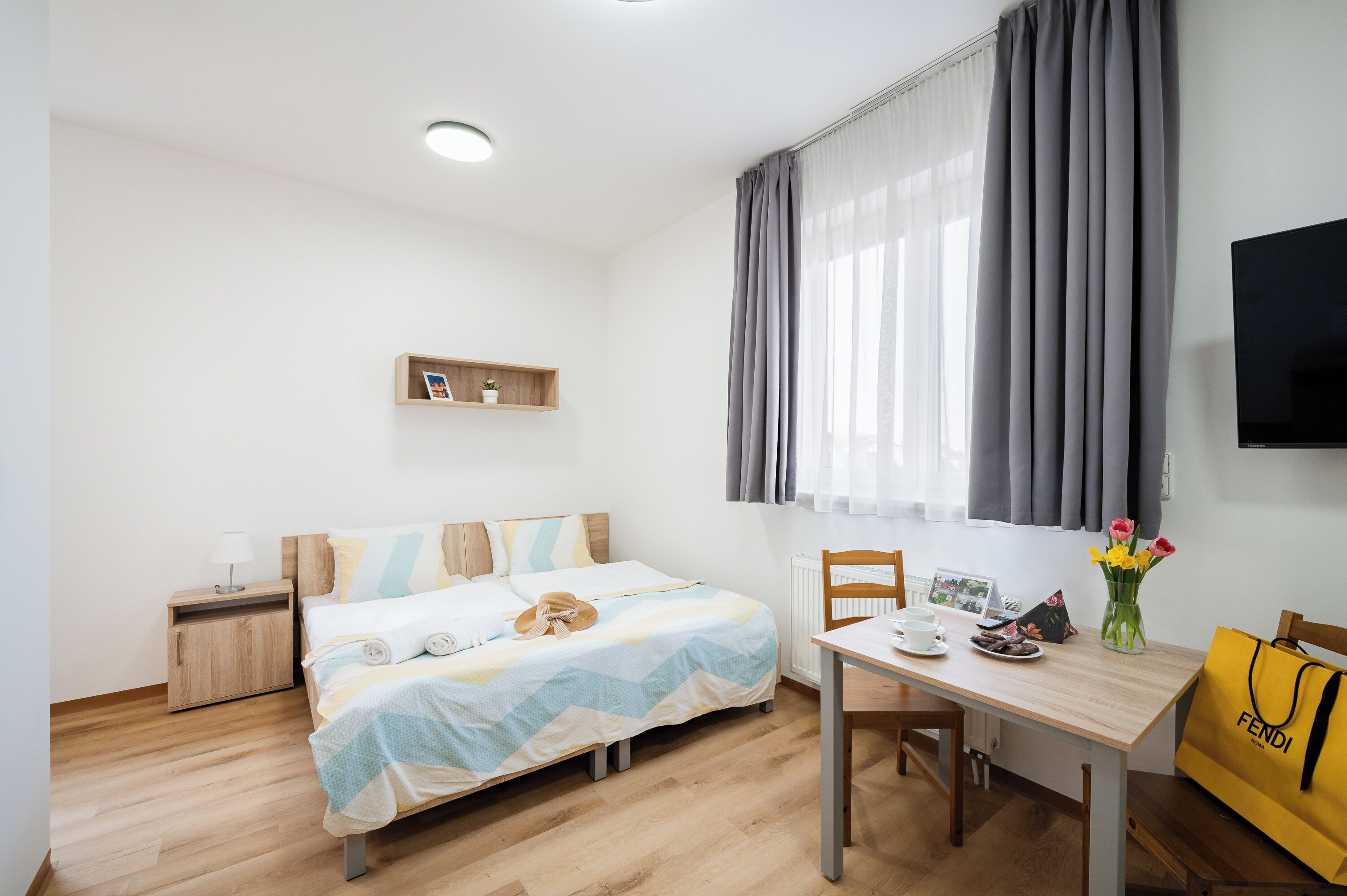 A neatly arranged hotel room with a bed, bedside table, a small shelf, a window with curtains, a TV mounted on the wall, and a dining table with a chair.