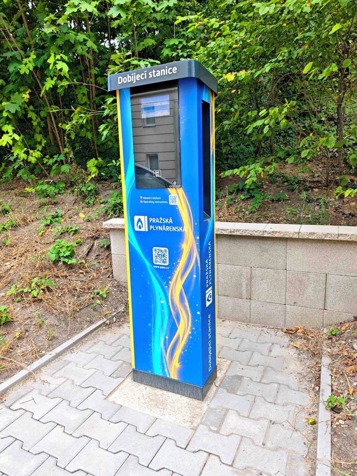 Quiosc blau i groc amb codi QR i un cartell que diu 'Dobijecí stanice' en un entorn de parc.