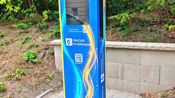 Quiosc blau i groc amb codi QR i un cartell que diu 'Dobijecí stanice' en un entorn de parc.