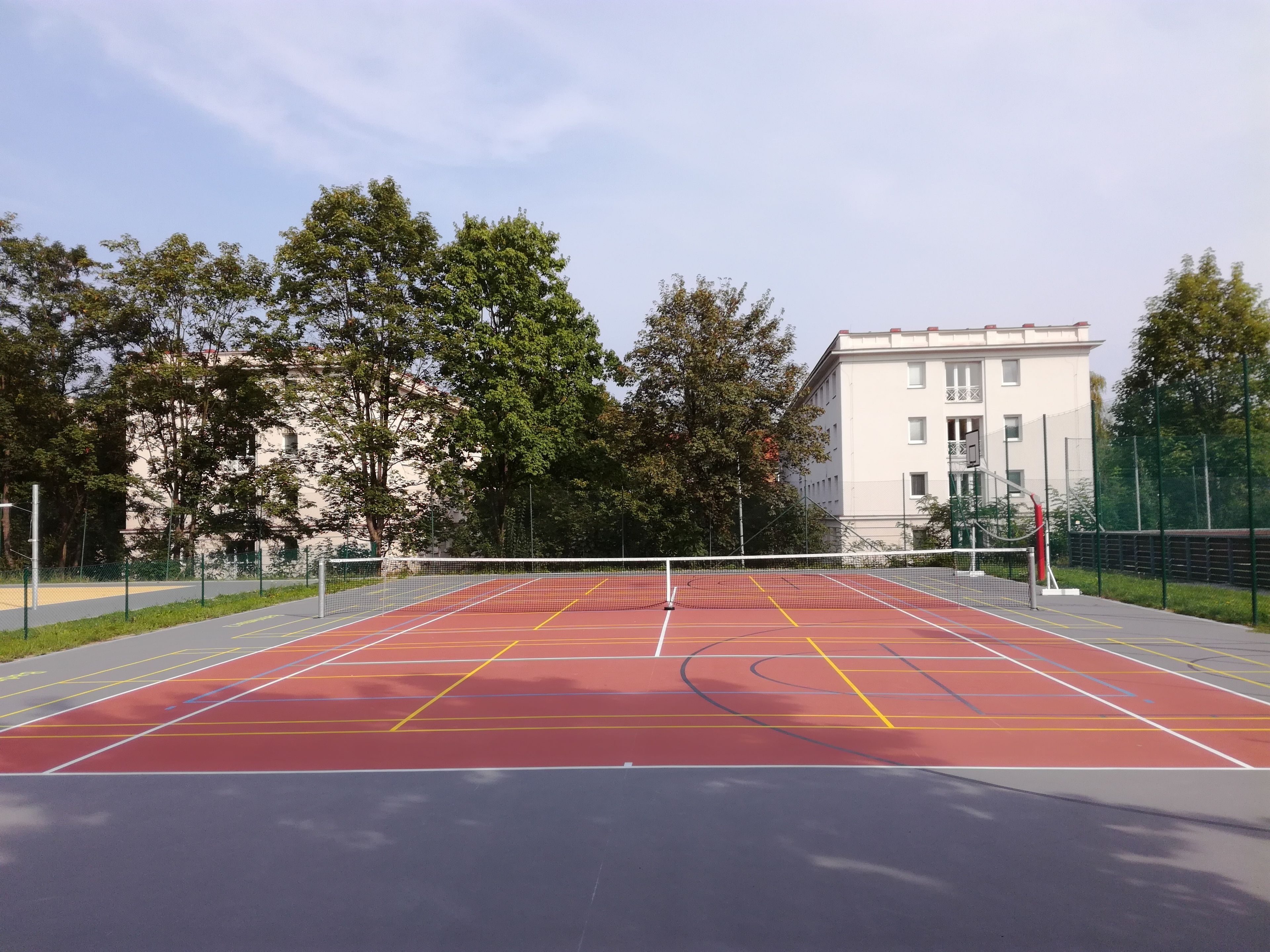 An outdoor tennis court with a net, surrounded by trees and a building in the background.