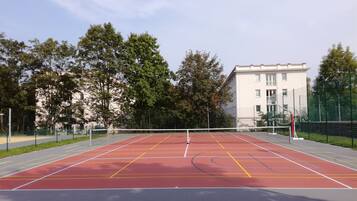An outdoor tennis court with a net, surrounded by trees and a building in the background.