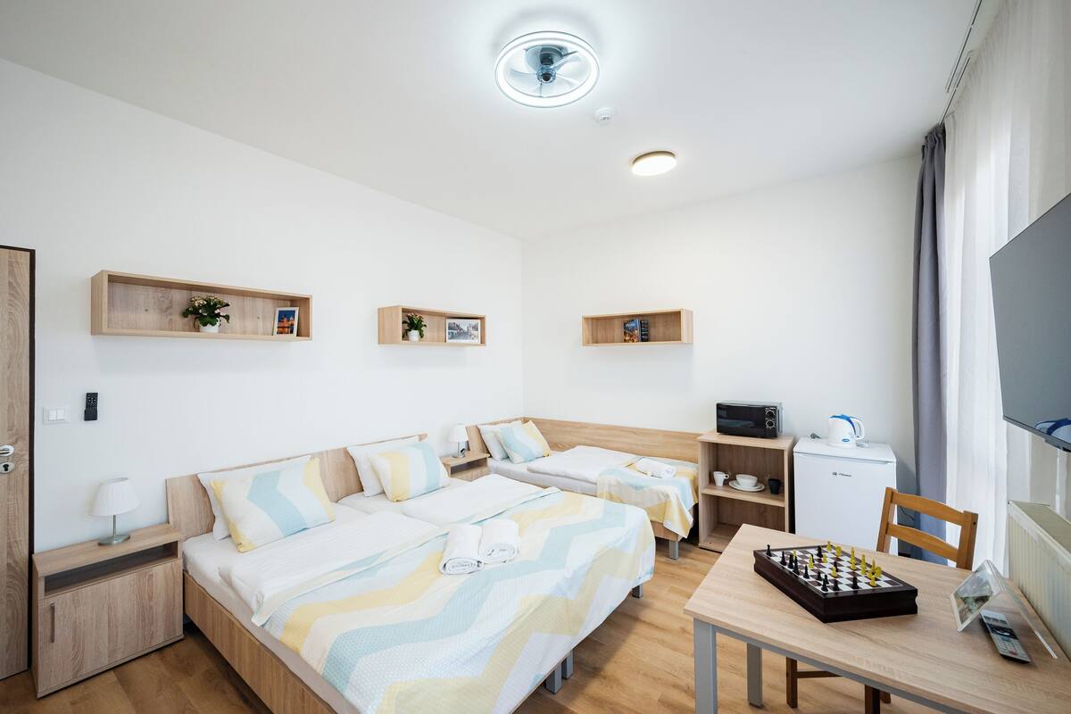 A hotel room with two beds, a desk with a chessboard, a TV, and a small kitchenette.