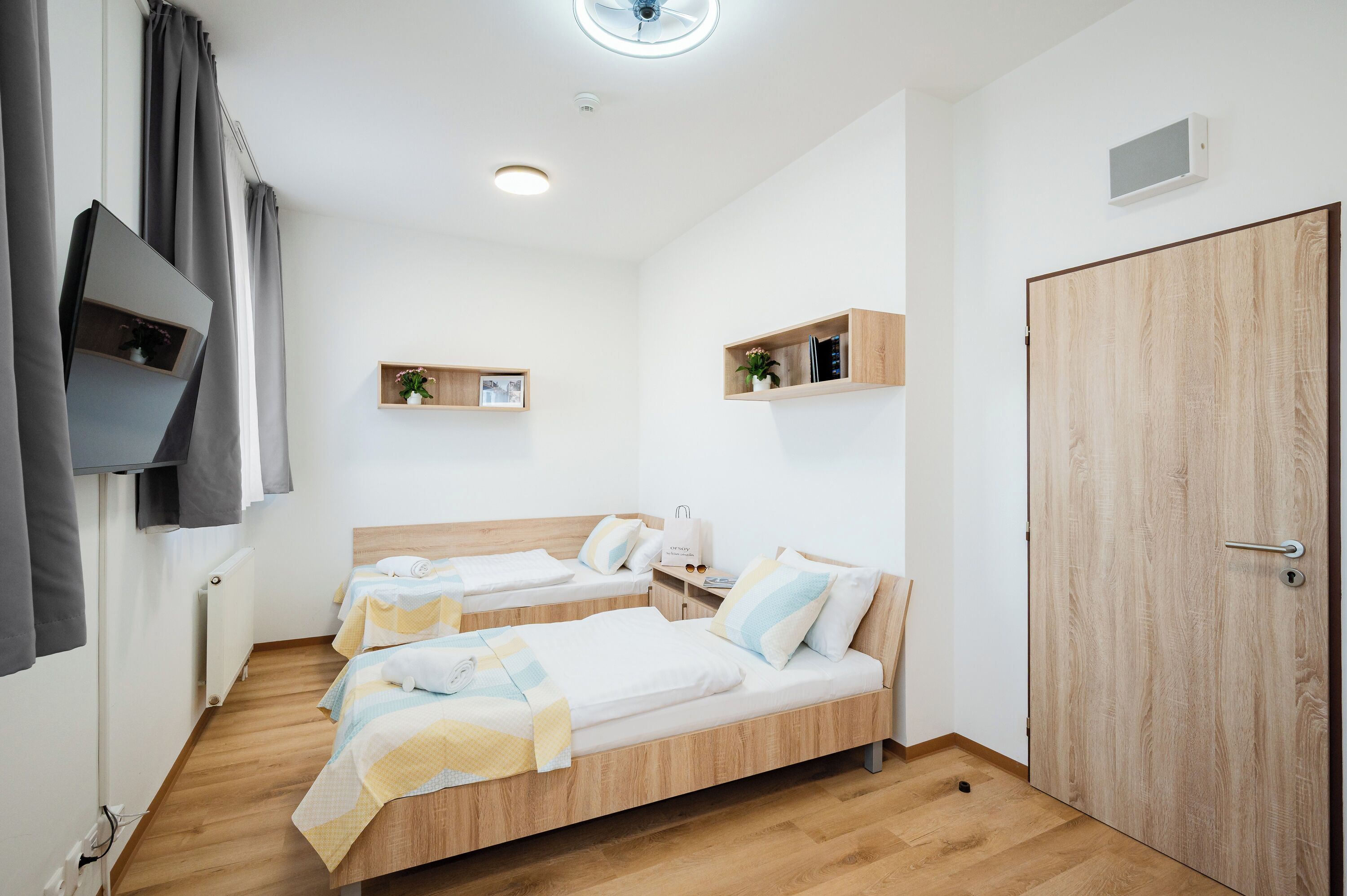A hotel room with two single beds, a wooden wardrobe, a small shelf with items, a television mounted on the wall, and a ceiling light.