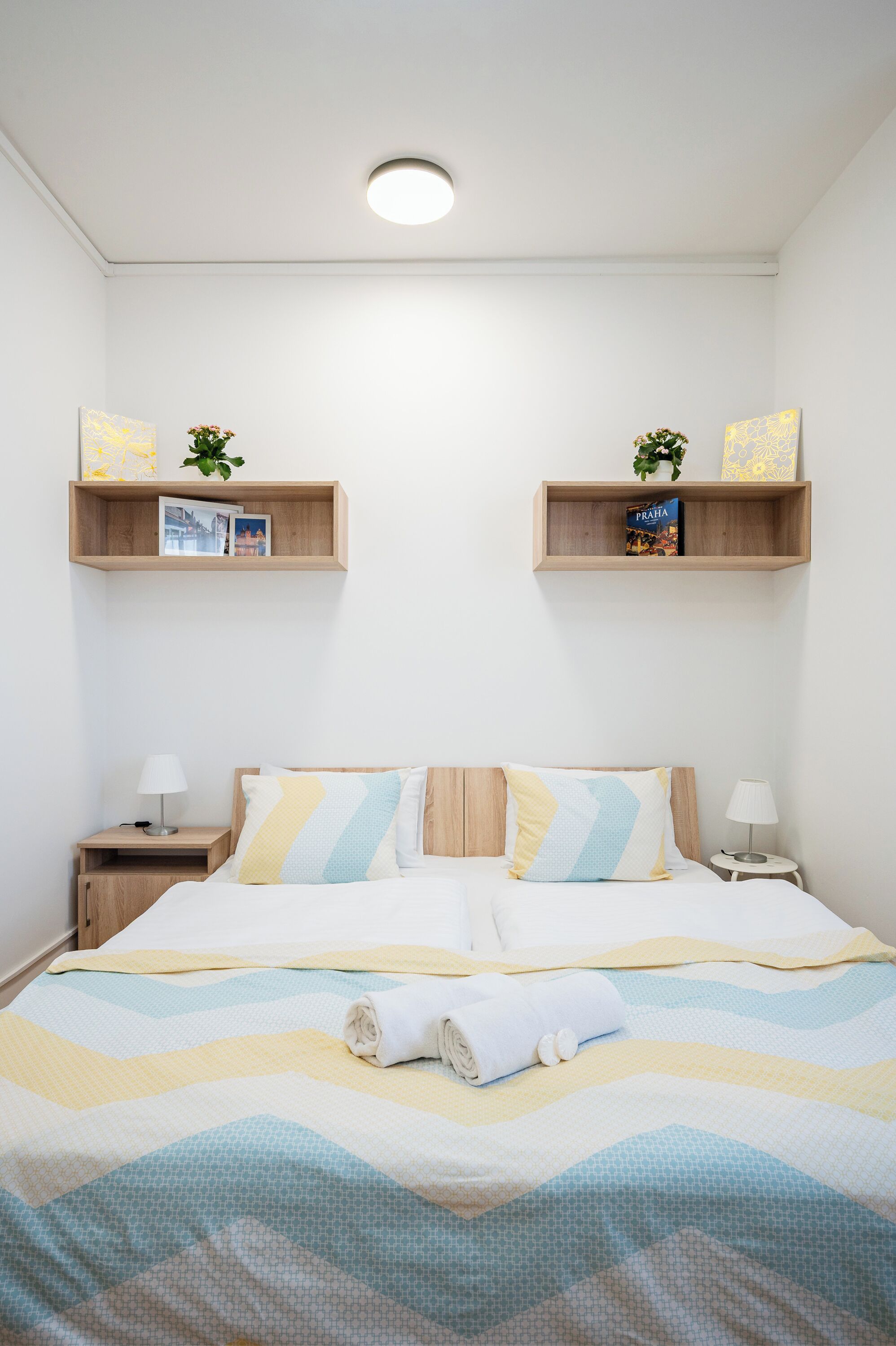 Une chambre d’hôtel bien rangée, avec un lit, des tables de chevet et des étagères murales ornées de livres et de plantes.
