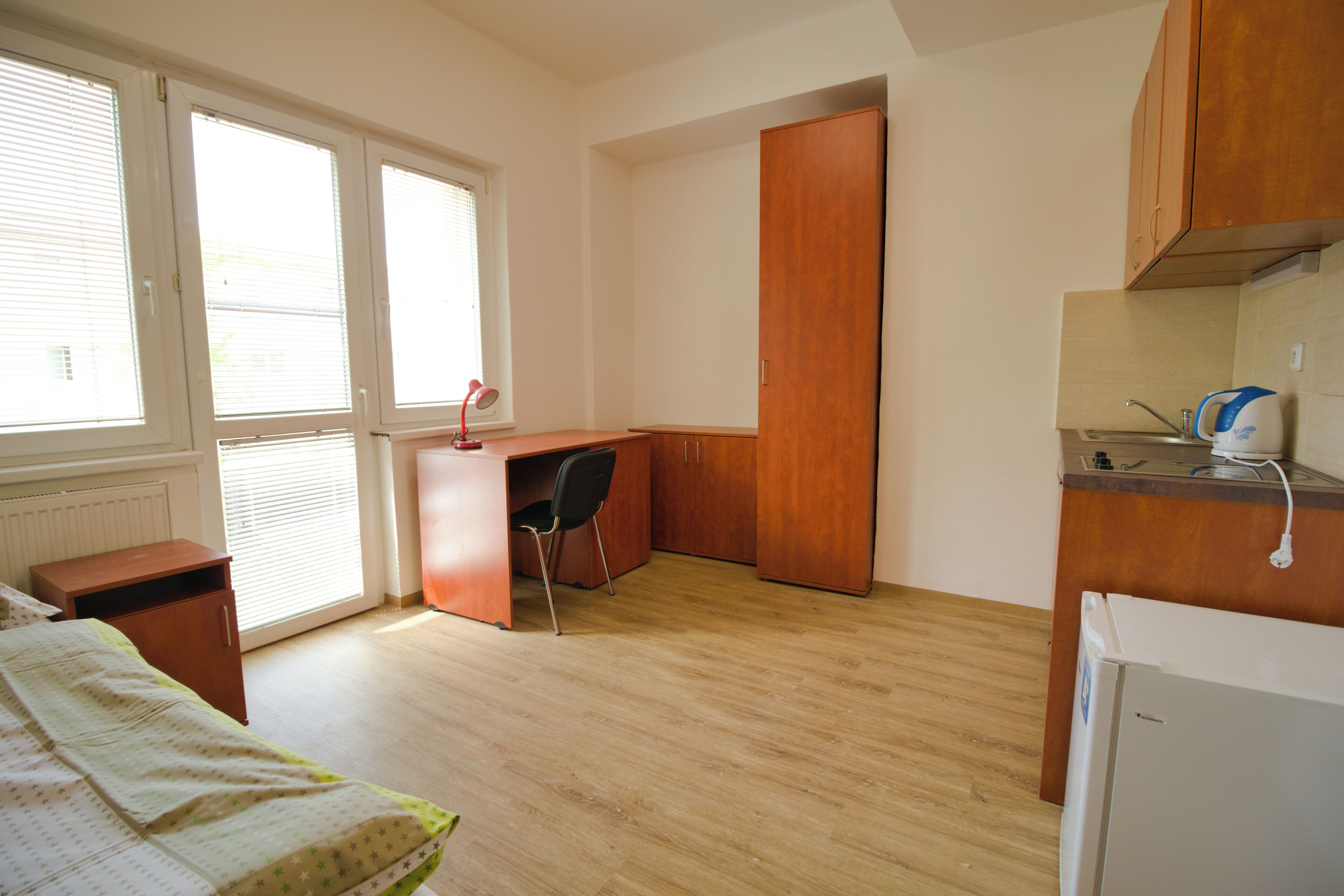 A bedroom with a bed, a desk, a chair, and a kitchenette.