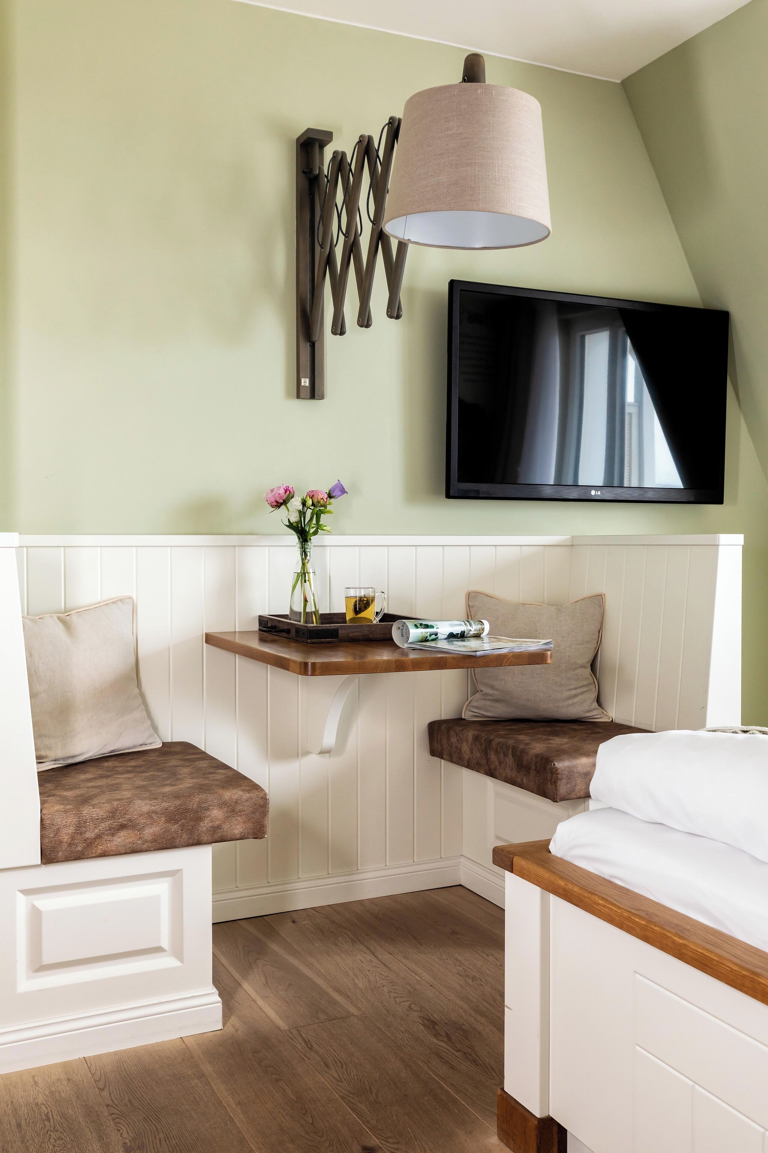 A room with a white bed, a bench with a cushion, a small table with a vase of flowers, a flat-screen TV mounted on the wall, and a pendant light.