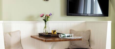 A room with a white bed, a bench with a cushion, a small table with a vase of flowers, a flat-screen TV mounted on the wall, and a pendant light.