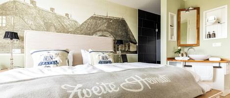 A hotel room with a bed featuring a travel-themed headboard, a bedside table with a lamp, a large mirror above the sink, and a wall with a black and white patterned mural.