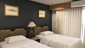 A hotel room with two beds, a nightstand with a lamp, framed pictures on the wall, and an air conditioning unit. - The 92 residence (Bangkok)