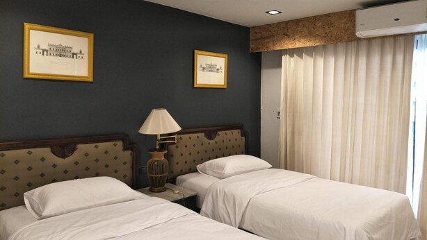 A hotel room with two beds, a nightstand with a lamp, framed pictures on the wall, and an air conditioning unit. - The 92 residence (Bangkok)