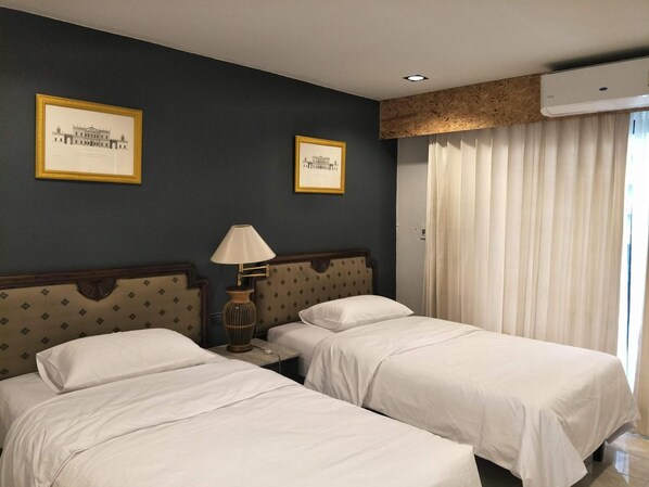 A hotel room with two beds, a nightstand with a lamp, framed pictures on the wall, and an air conditioning unit. - The 92 residence (Bangkok)