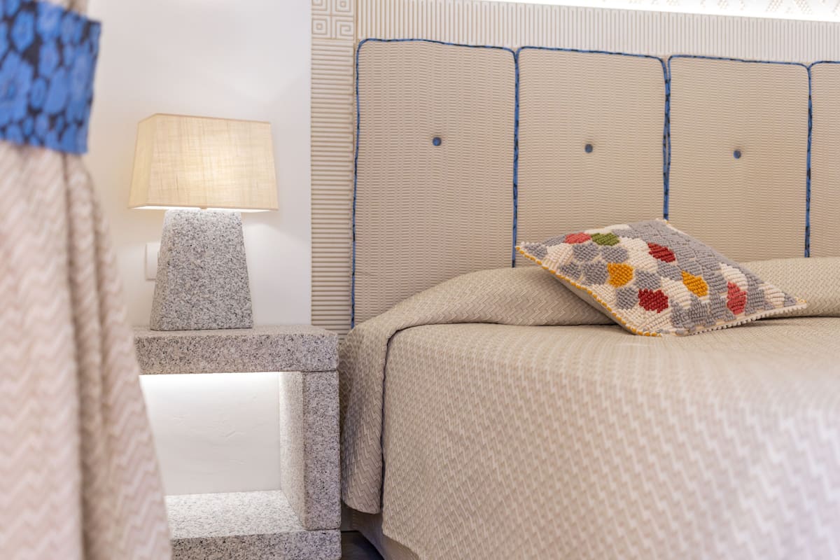 A neatly made bed with a patterned pillow, a bedside lamp, and a textured headboard.