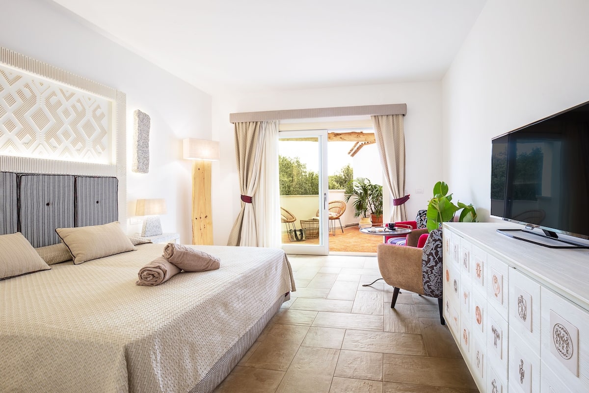 A hotel room with a bed, a TV, a cabinet with drawers, a balcony with a table and chairs, and a view of greenery outside.