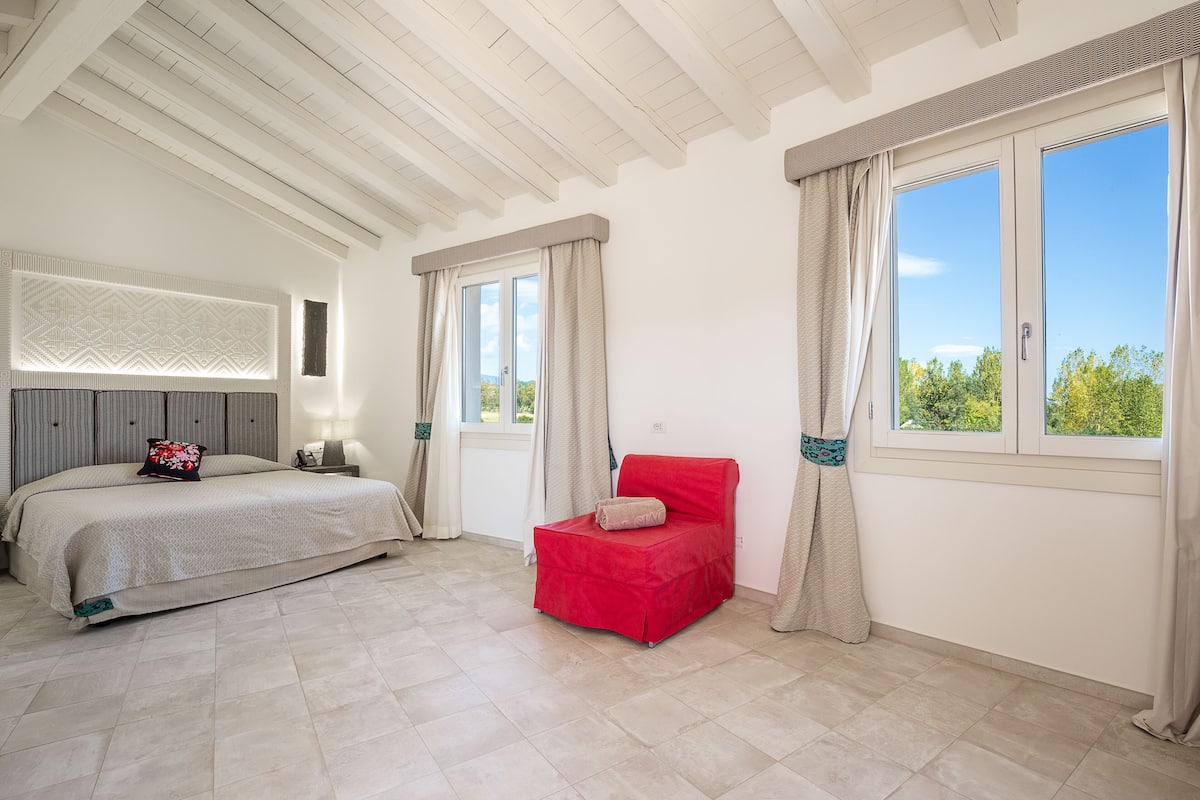 A bedroom with a bed, a red chair, a window with curtains, and a view of greenery outside.