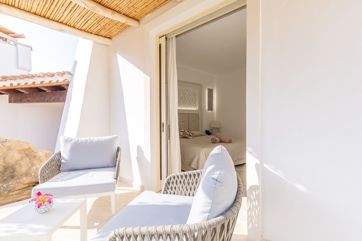 A balcony with wicker chairs and a table, a bed with a patterned headboard, and a view of the building's exterior.