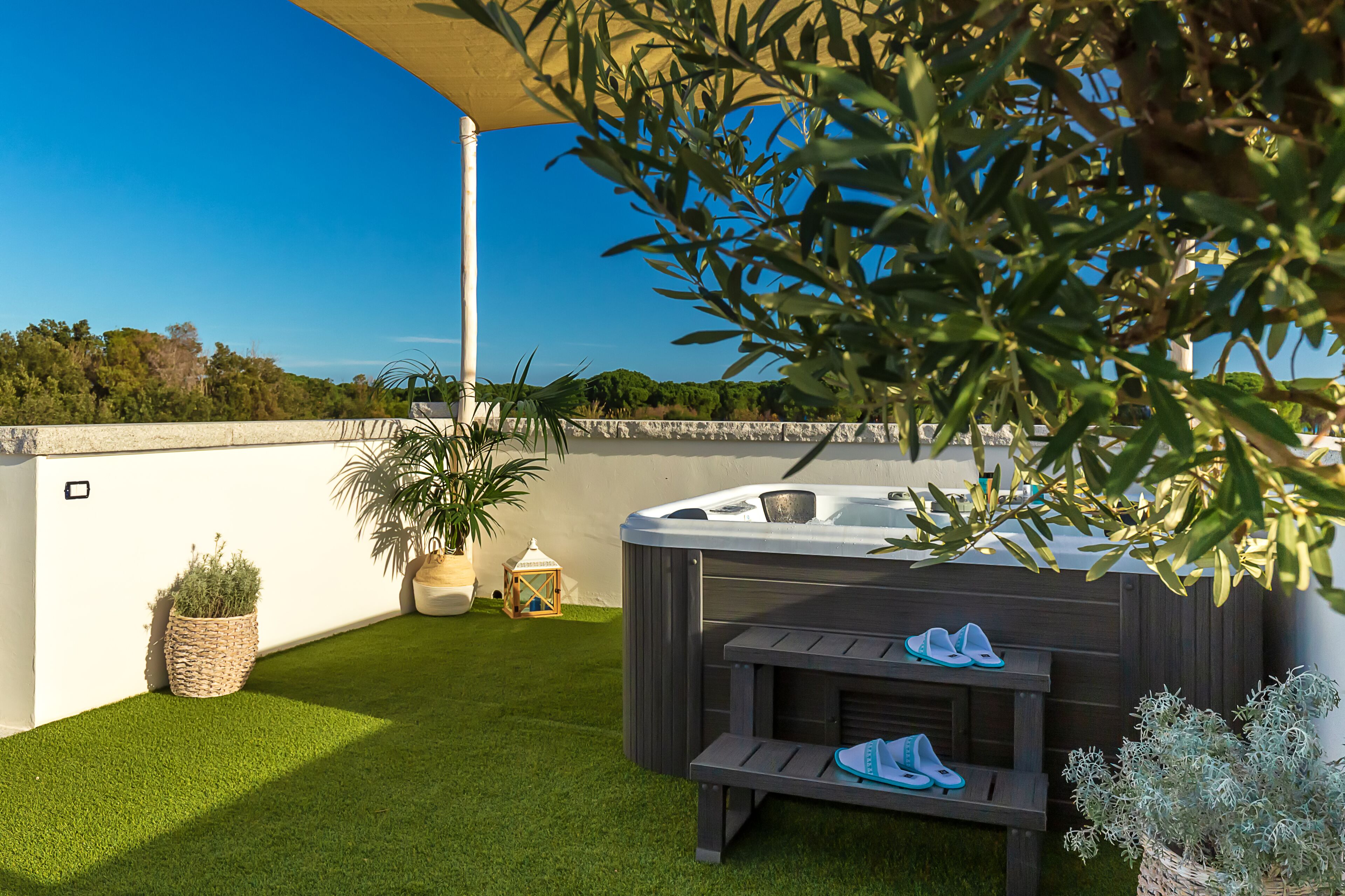 Een hottub met een houten bankje en een paar slippers, omringd door een verzorgd gazon en potplanten.