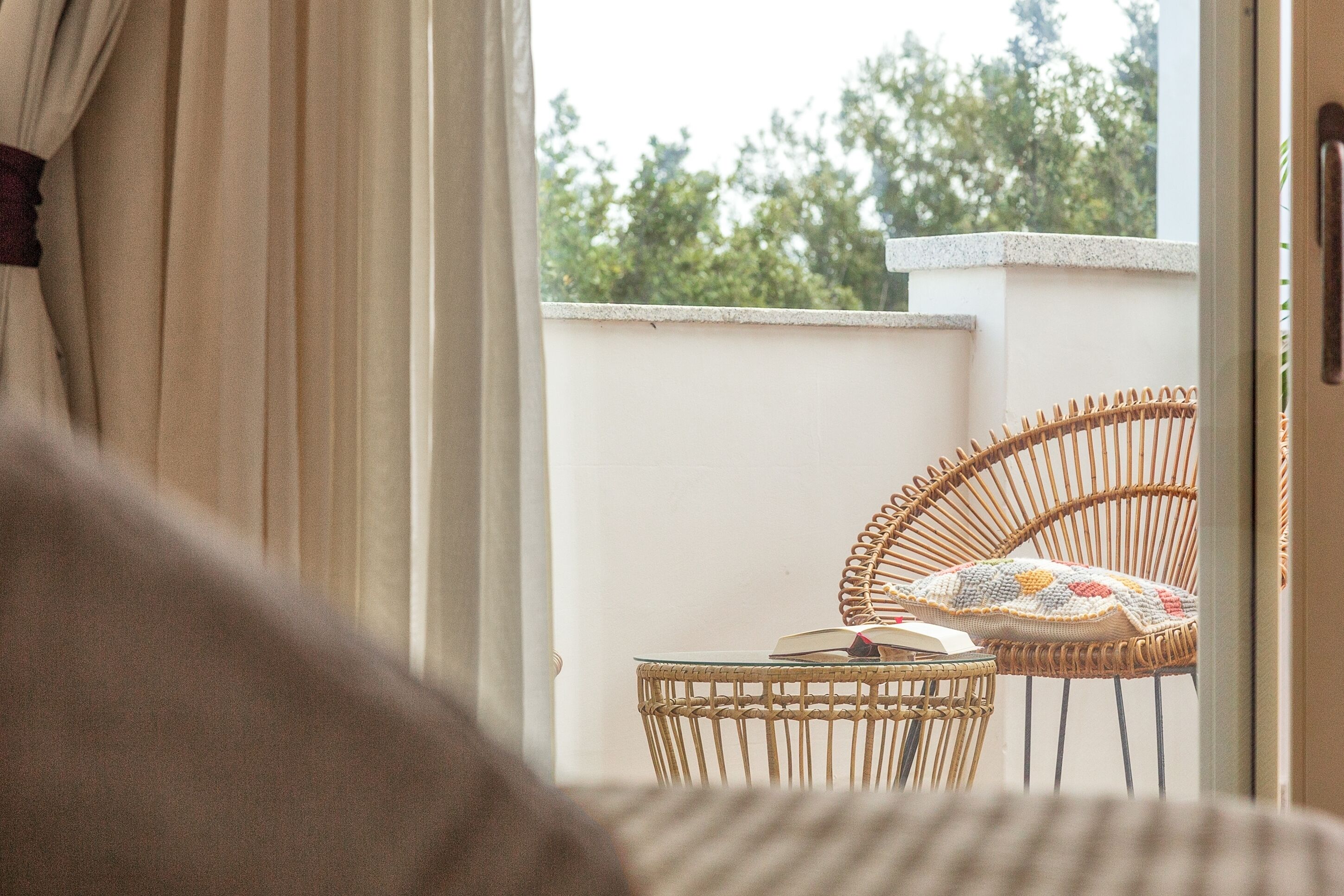 A wicker chair with a cushion and a book on a small table, positioned near a window with curtains.