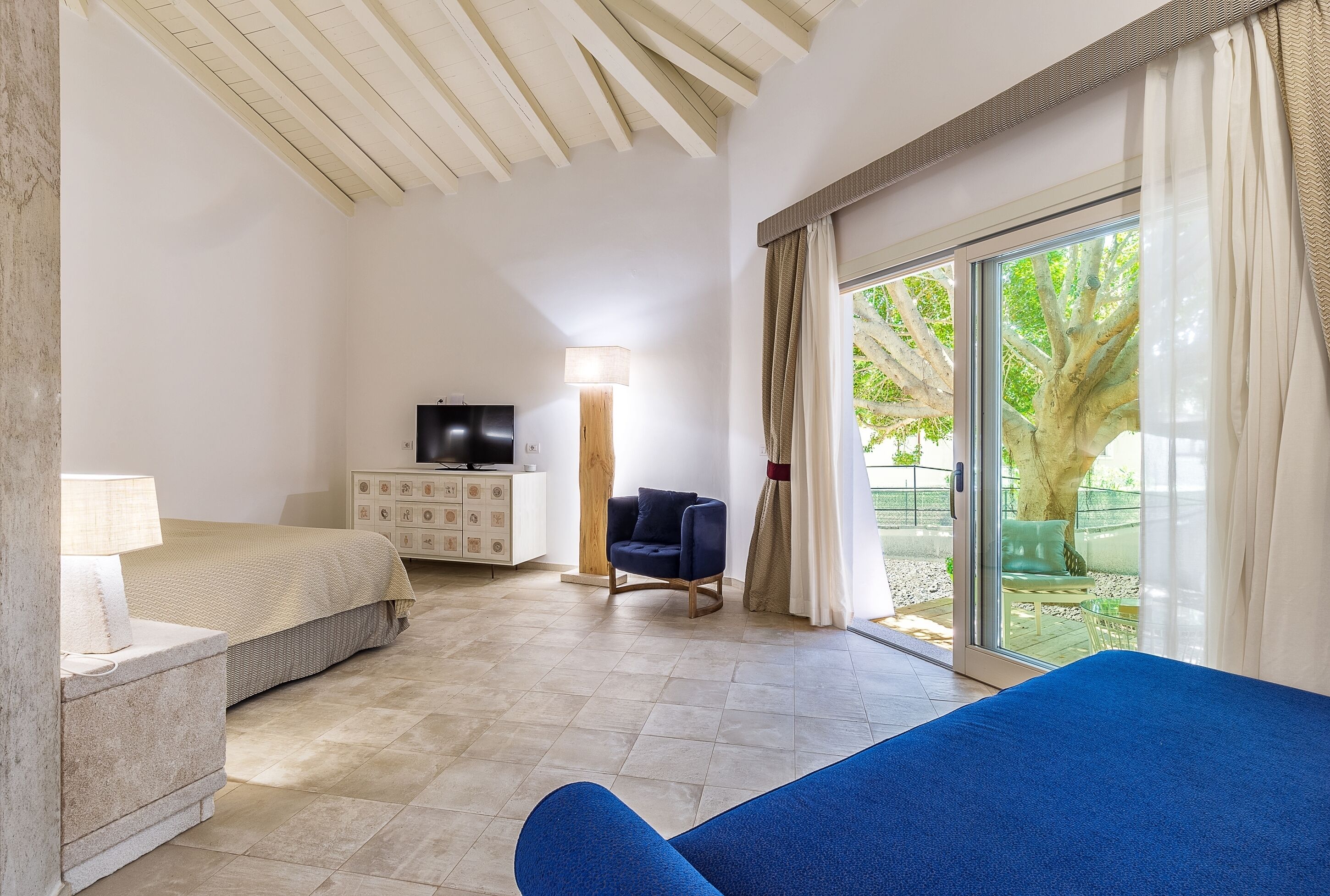 Un dormitorio con cama, escritorio, silla, televisor y un ventanal con vistas a zonas verdes.