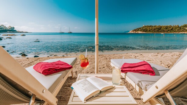 Une vue sur la plage avec deux chaises longues, un parasol, une boisson et un livre posés sur le sable.