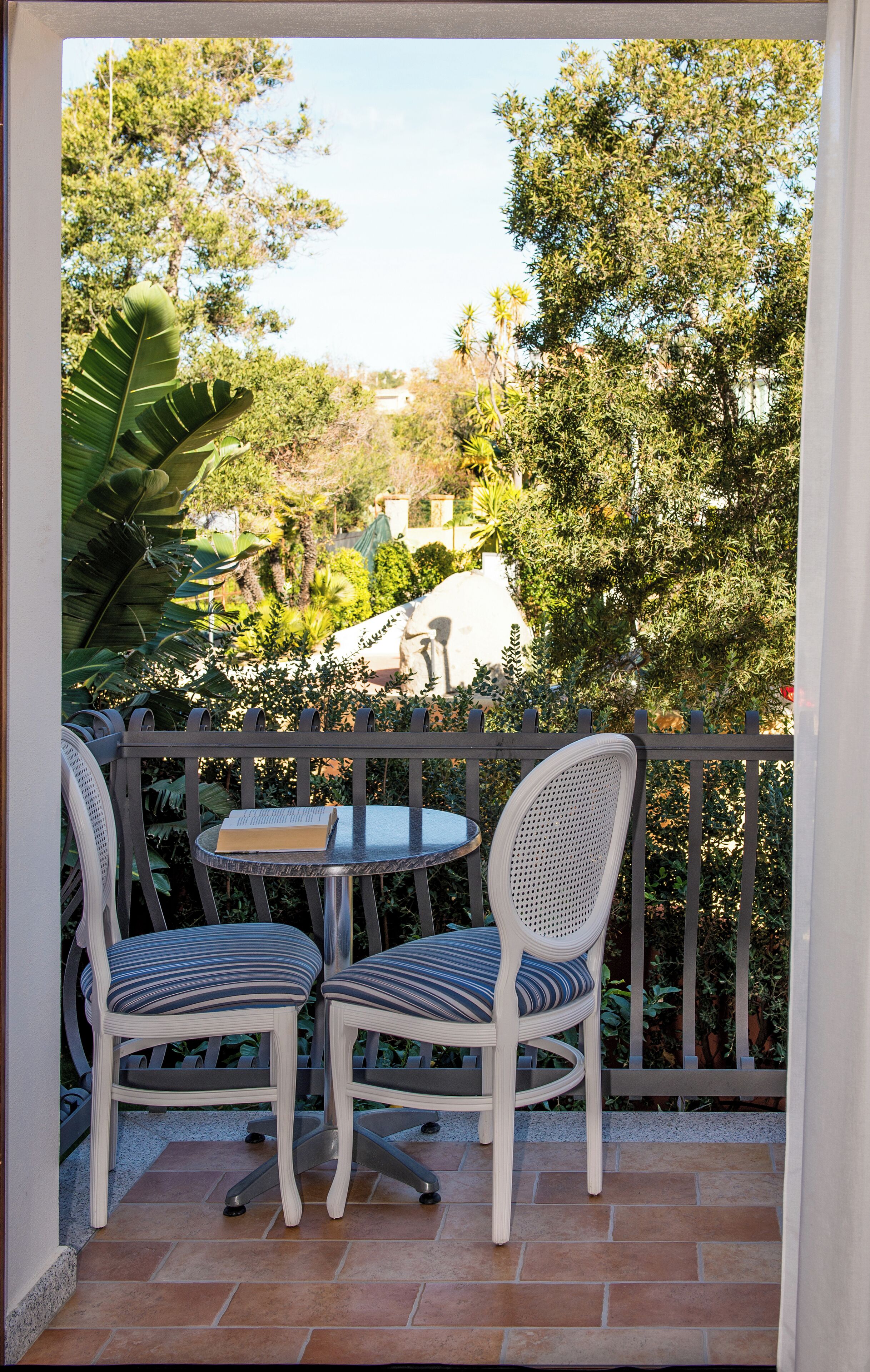 A balcony with two white chairs and a small table, overlooking a garden with various plants and a white building in the background.