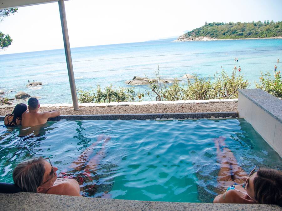 A pool with clear blue water, surrounded by a stone wall, overlooking a beach with people swimming.