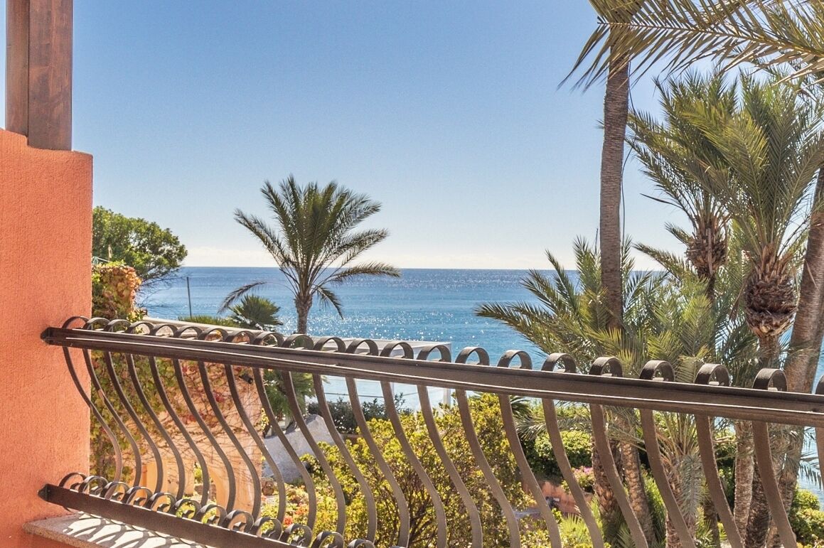 Ein Balkon mit schmiedeeisernem Geländer, der auf eine Küstenlandschaft mit Palmen und Meer blickt.