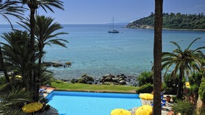 Une piscine bordée de parasols jaunes, de palmiers, et offrant une vue sur la mer et les bateaux.