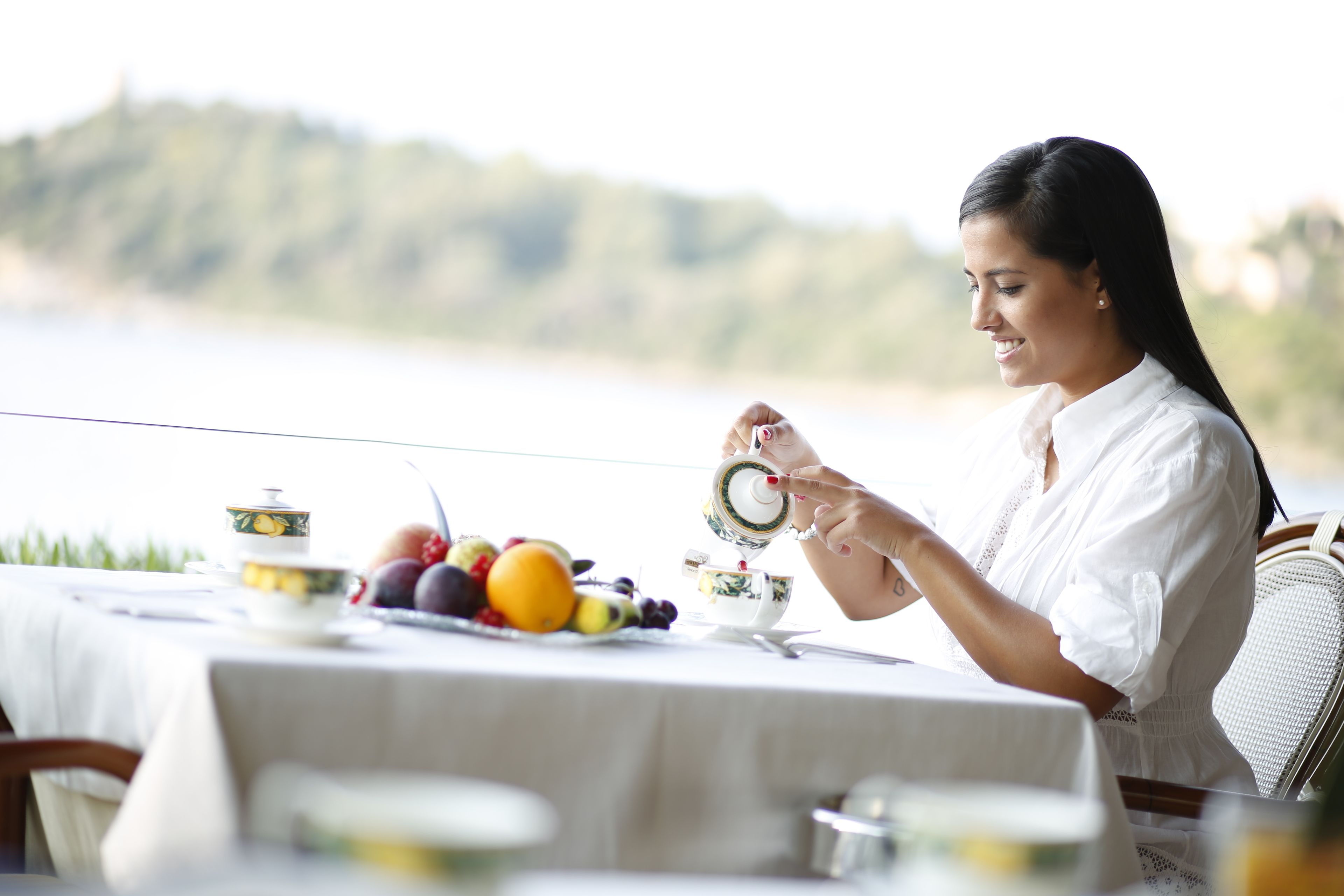 A dining table with a white tablecloth, a teapot, cups, and a variety of fruits.