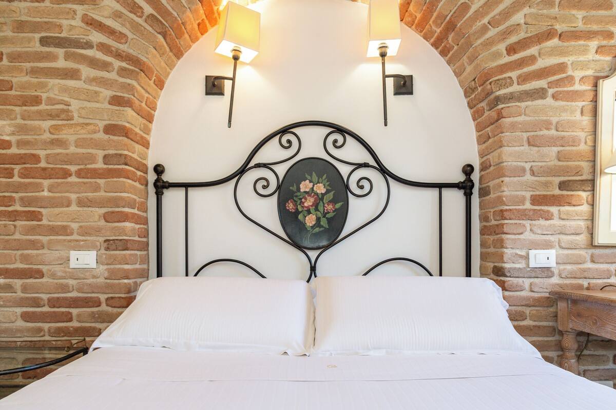 Un lit avec du linge de lit blanc, dans une pièce aux murs en briques et dotée d’une tête de lit décorative en métal.