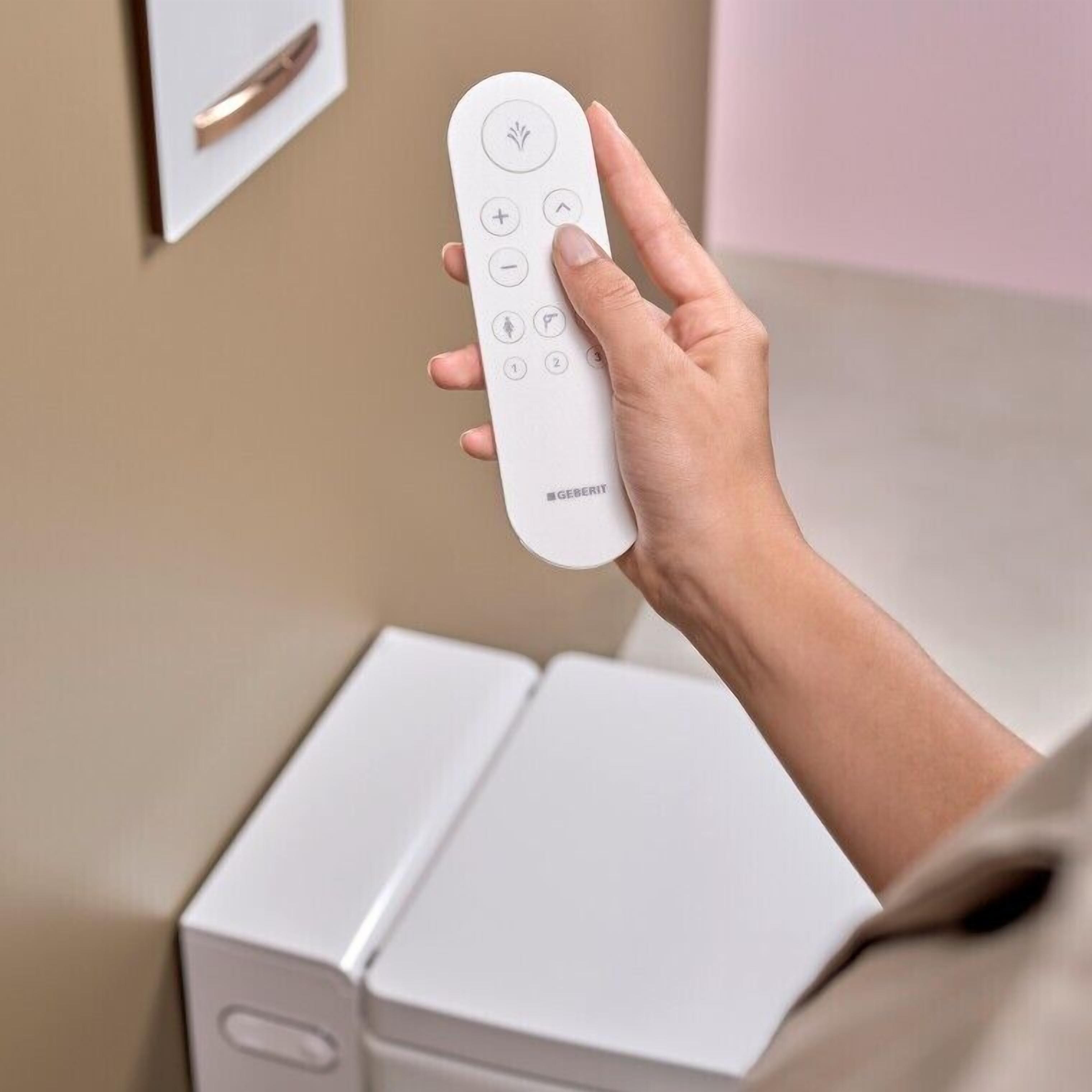 Una mano sostiene un control remoto de diseño minimalista, sobre una superficie blanca con una pared beige al fondo.