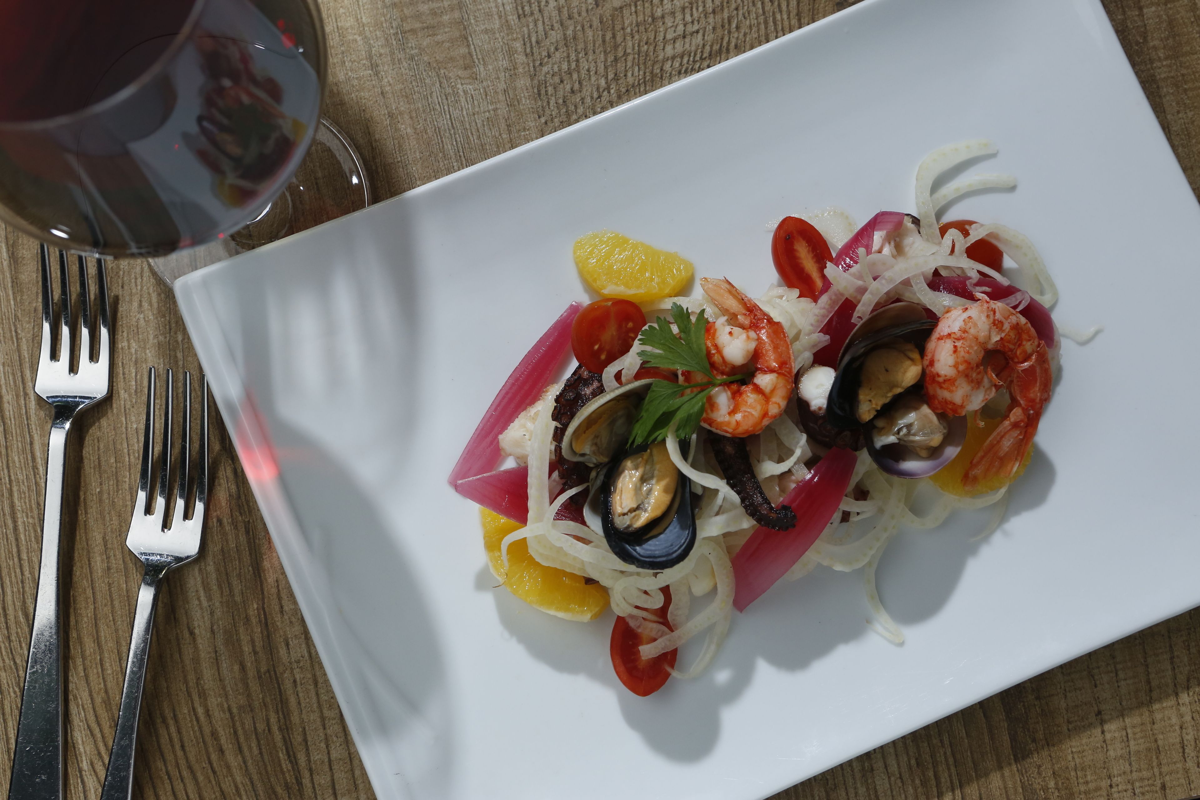 Un plato blanco con un guiso de mariscos, un tenedor y una copa de vino tinto sobre una mesa de madera.