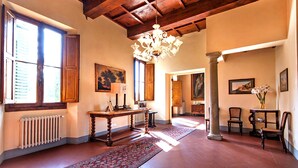 A spacious room with a high wooden ceiling, a chandelier, a large window with shutters, a table with a lamp, and a chair with a vase of flowers.
