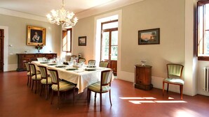 A dining room with a long table set for six, surrounded by six chairs. There is a chandelier hanging from the ceiling, and the room has large windows allowing natural light.