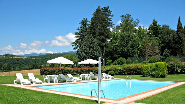 A rectangular swimming pool with a stone border, surrounded by white lounge chairs and umbrellas, set against a backdrop of lush greenery and distant mountains.