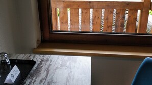 Une pièce avec une fenêtre donnant sur une terrasse en bois et sur de la verdure. On aperçoit une table avec un plateau et une chaise.