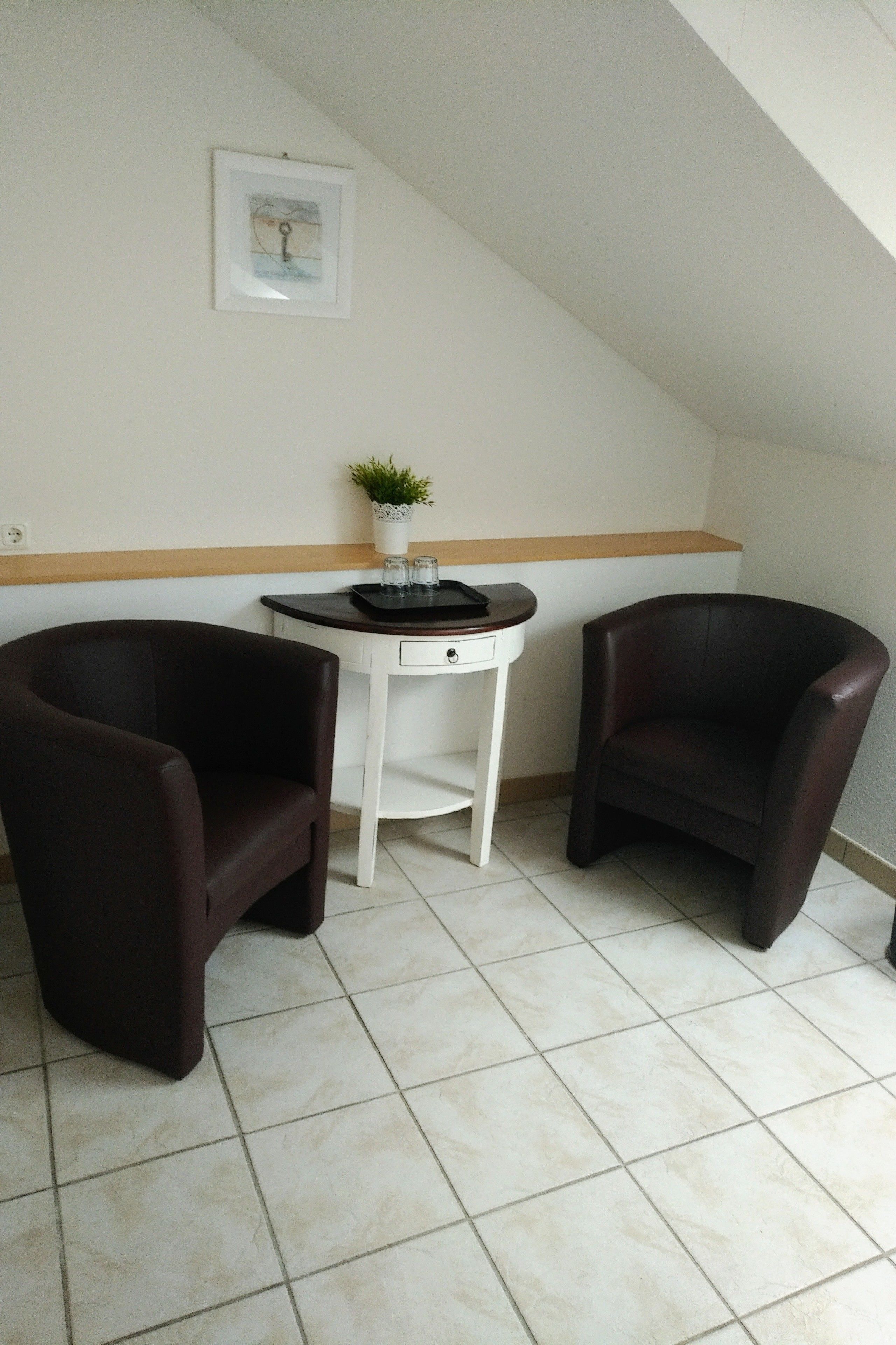 Um quarto pequeno com duas cadeiras marrom-escuro, uma mesa branca com um vaso de planta e um quadro na parede.