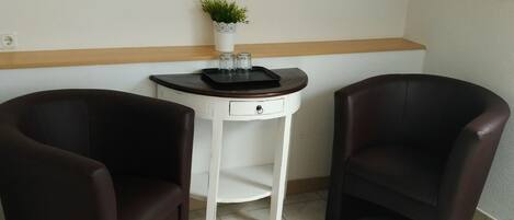 Um quarto pequeno com duas cadeiras marrom-escuro, uma mesa branca com um vaso de planta e um quadro na parede.