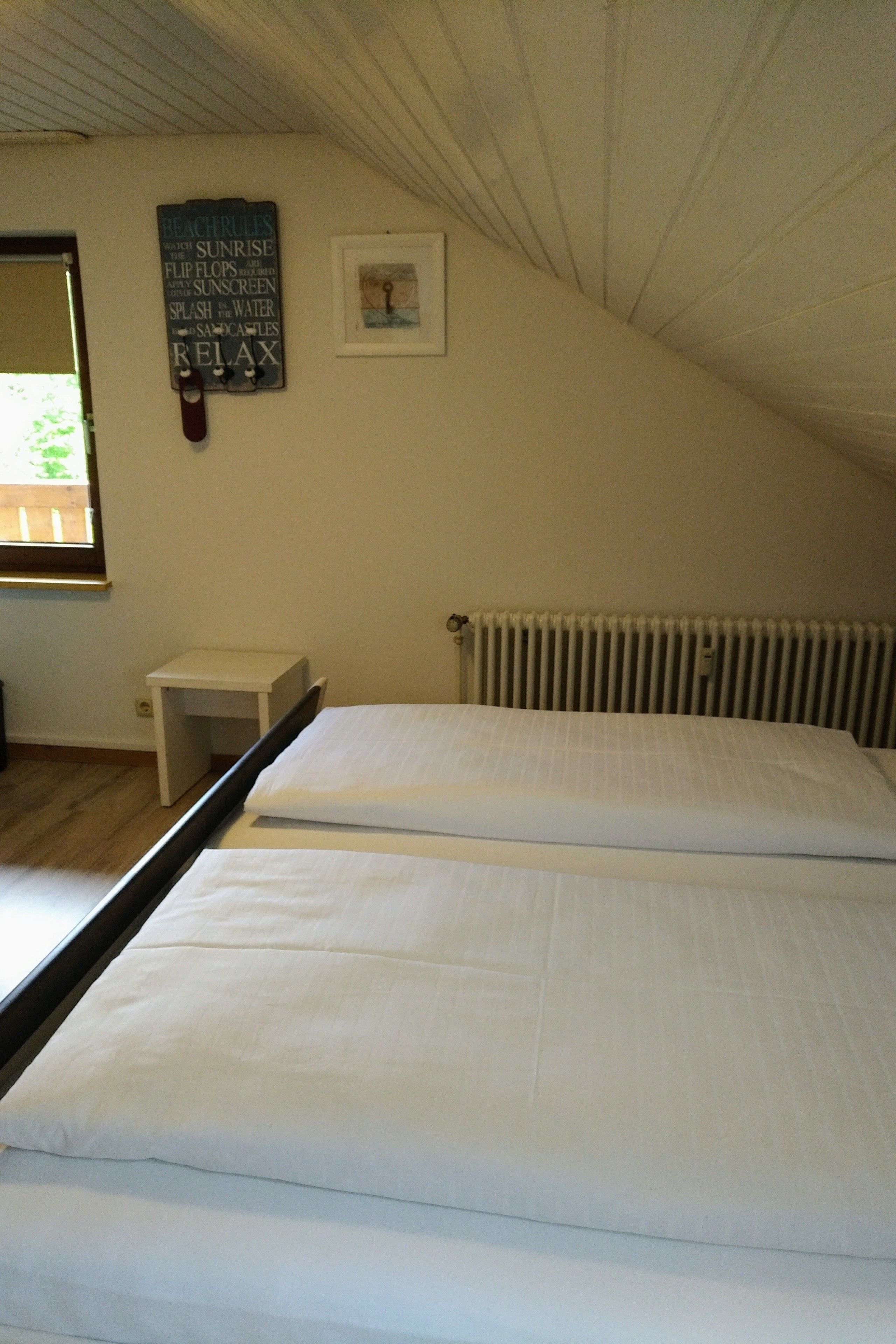 A bedroom with a bed, a small bedside table, a radiator, a window, and a wall with a sign.