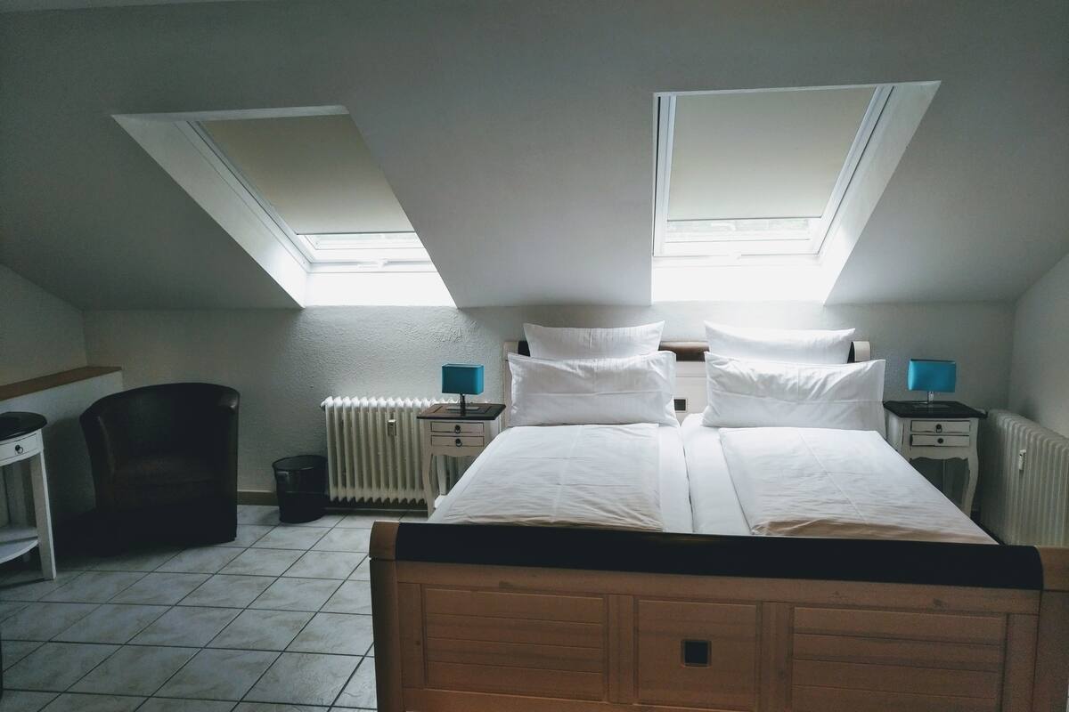 Un dormitorio con techo inclinado, tragaluces, cama de madera, ropa de cama blanca, dos mesitas de noche con lámparas, un radiador y una silla.