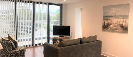 A modern living room with a grey sofa, a patterned armchair, a flat-screen TV, and a large window with blinds.