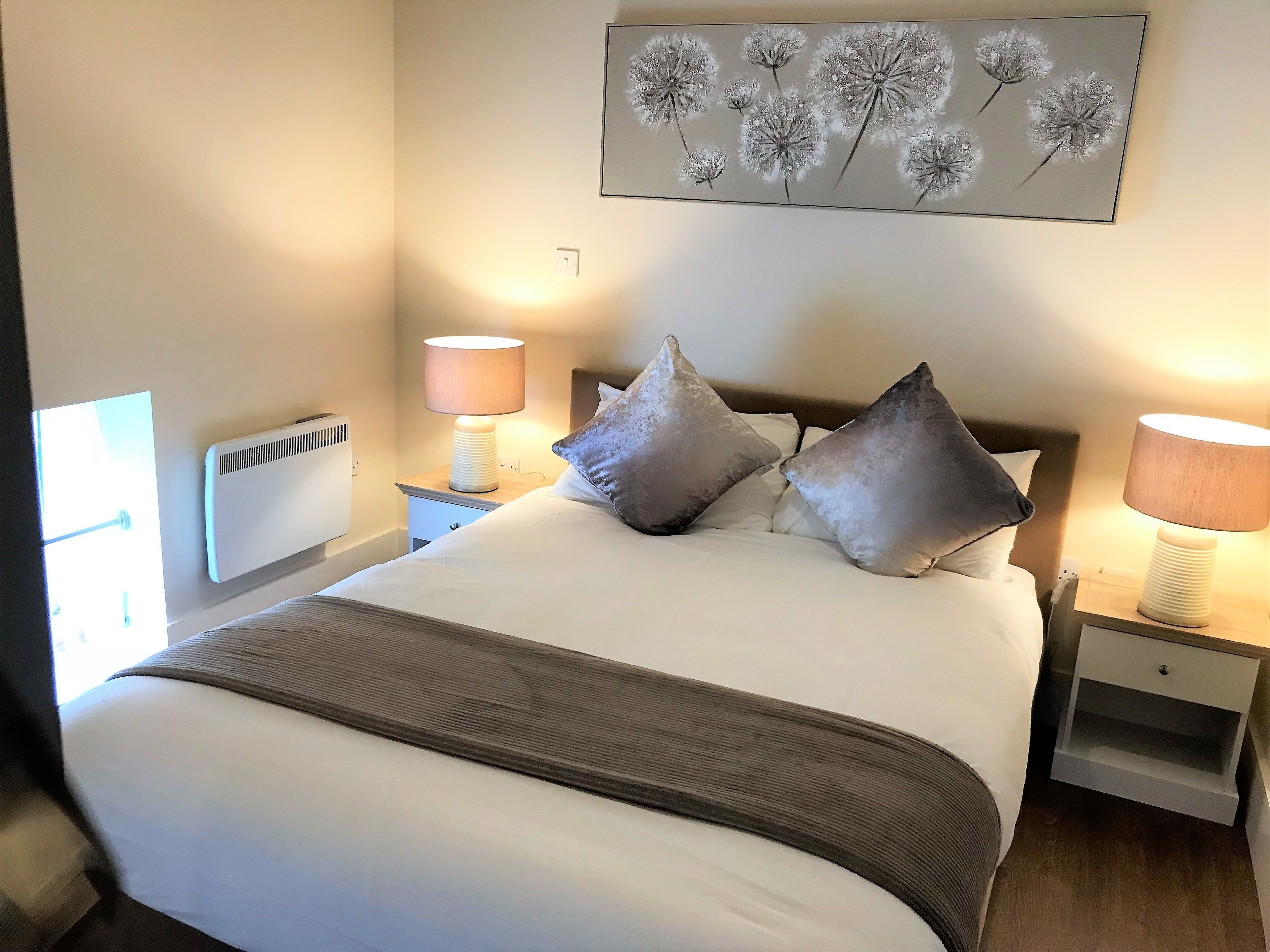 A neatly made bed with pillows, a bedside lamp, and a wall-mounted artwork of dandelions.