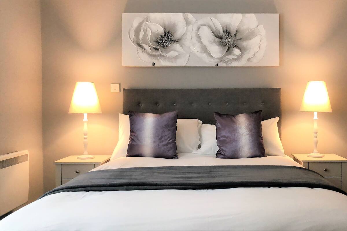A neatly made bed with a dark headboard, two bedside lamps, and a large floral artwork above the bed.