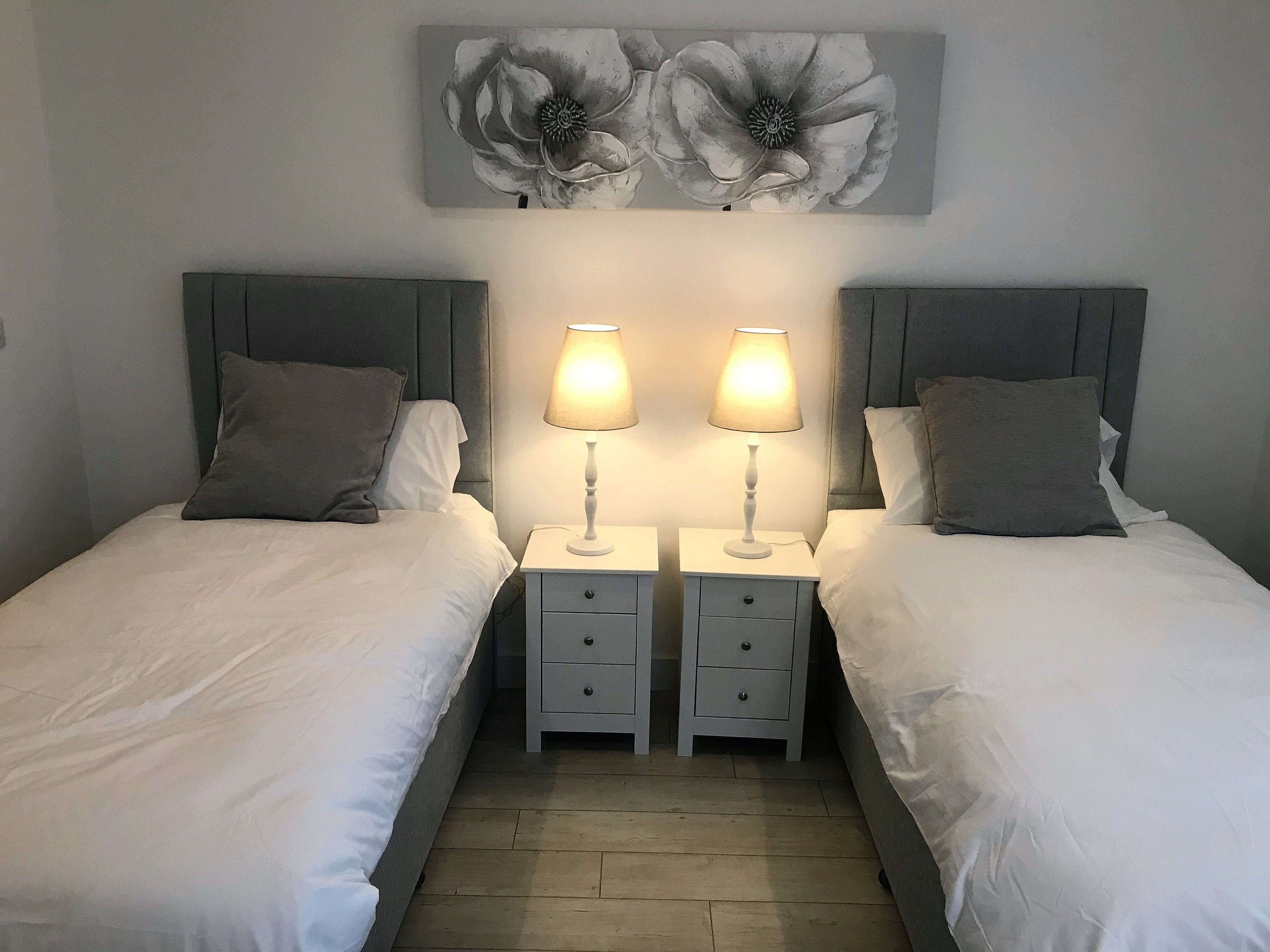 A twin bedroom with two beds, each with a grey headboard, white bedding, and a bedside table with a lamp. There is a large floral artwork above the beds.