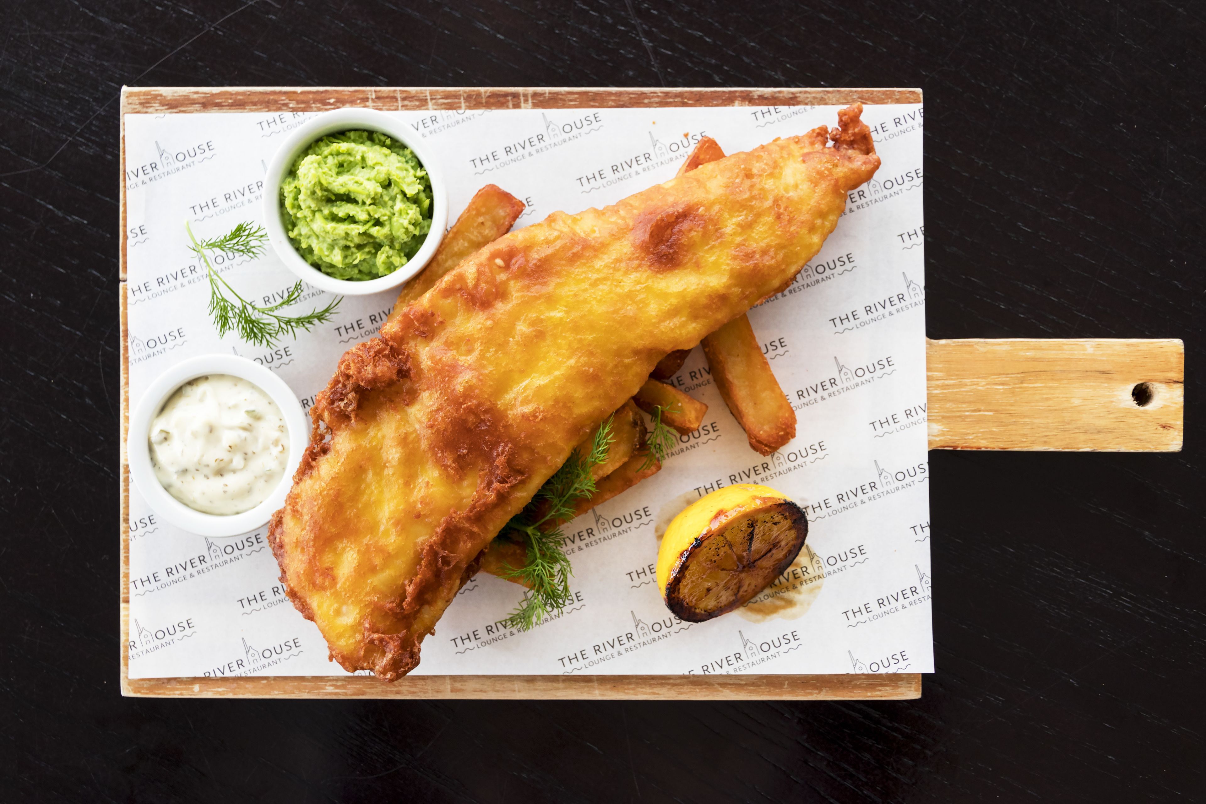 A fish and chips meal with sides of mushy peas and tartar sauce on a branded serving board.