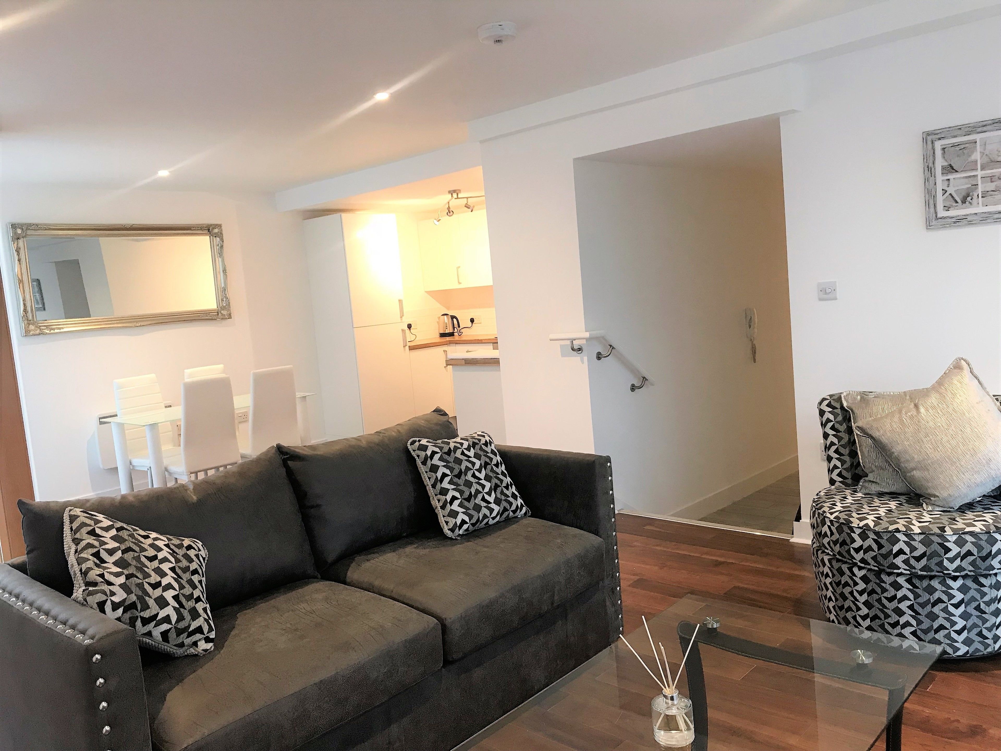 A modern living room with a grey sofa, patterned cushions, a glass coffee table, and a patterned ottoman.